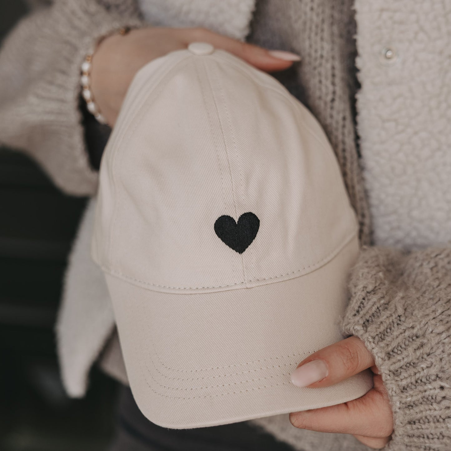 Beige Baseball Cap mit schwarzem Herz-Stick in Händen gehalten