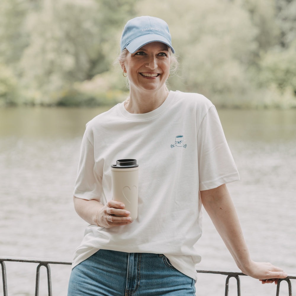 Frau mit weißem Coffee Club T-Shirt und Becher am Wasser, entspannter Lifestyle