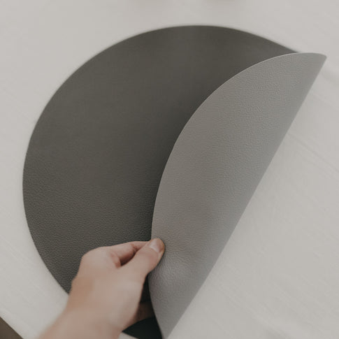 Hand dreht ein rundes, graues, wendbares Platzset mit strukturierter Oberfläche auf einem weißen Tisch um.