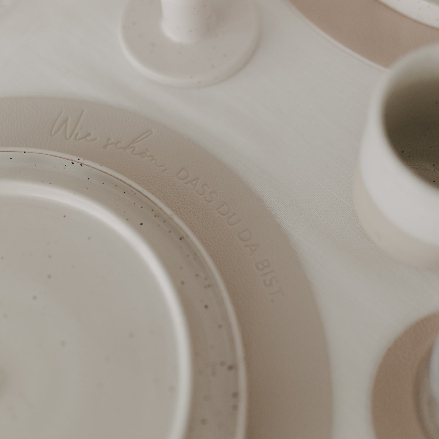 Cremefarbenes Platzset mit Prägung „Wie schön, dass du da bist“ und Keramikgeschirr auf hell gedecktem Tisch.