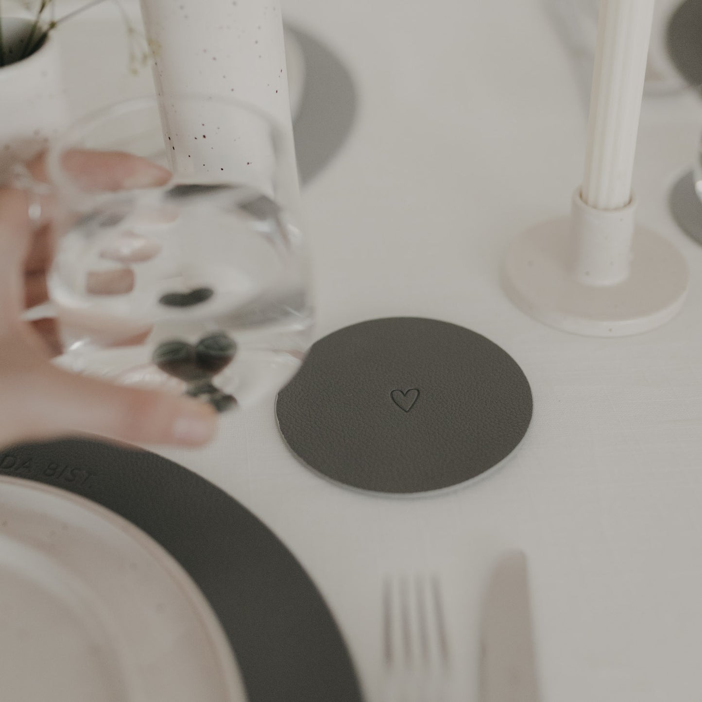 Dunkelgrauer Herz-Untersetzer auf weißem Tisch, gehalten neben Wasserglas.