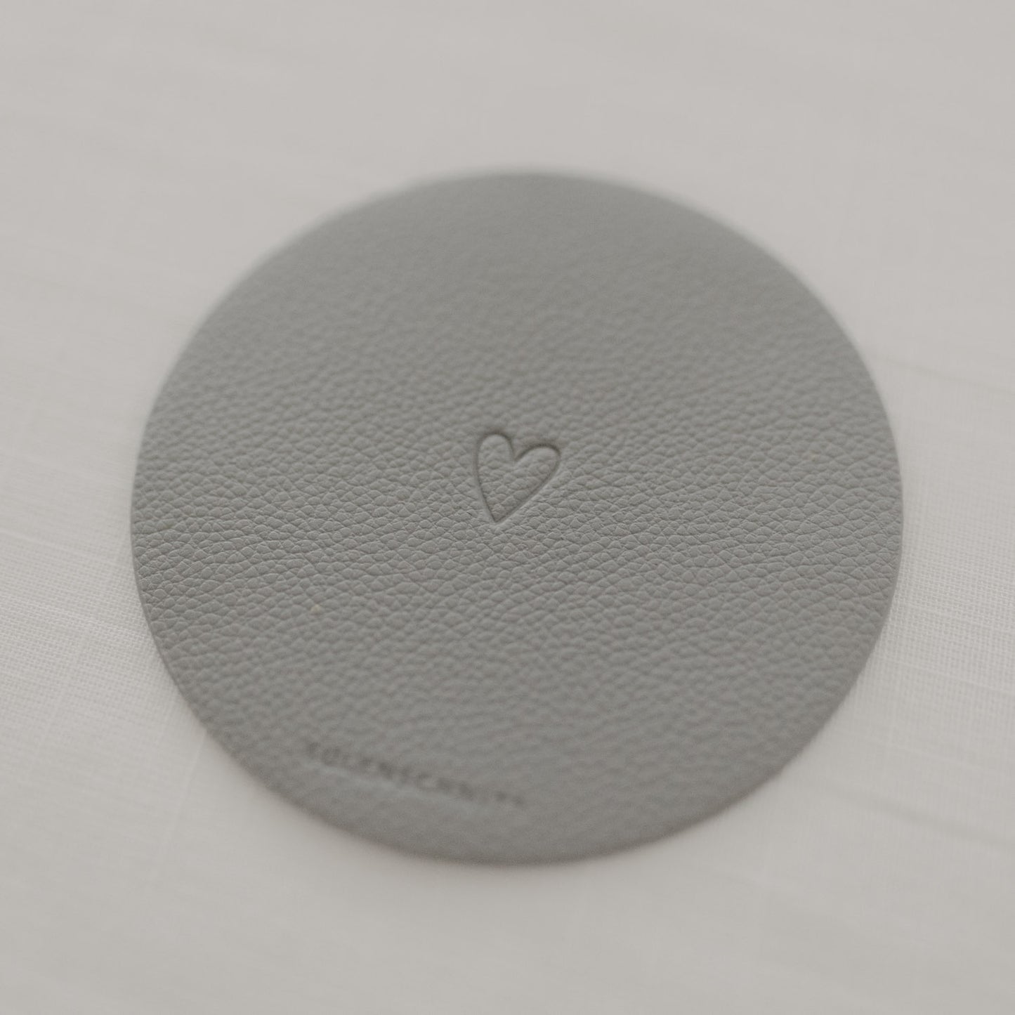 Hellgrauer Untersetzer mit Herzprägung und Eulenschnitt-Logo auf Tischdecke.