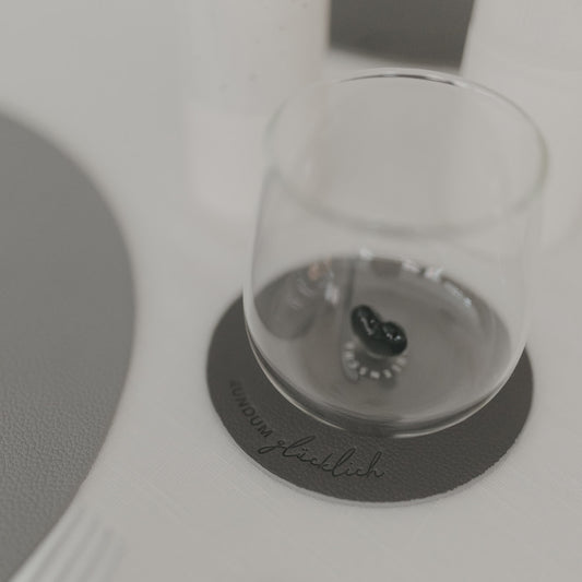 Grauer Untersetzer „Rundum glücklich“ mit Glas darauf, modernes Design auf hell gedecktem Tisch.