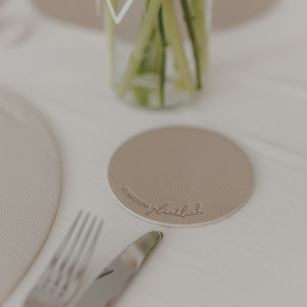 Untersetzer in Creme mit Schrift „Rundum glücklich“ auf gedecktem Tisch neben Vase und Besteck.