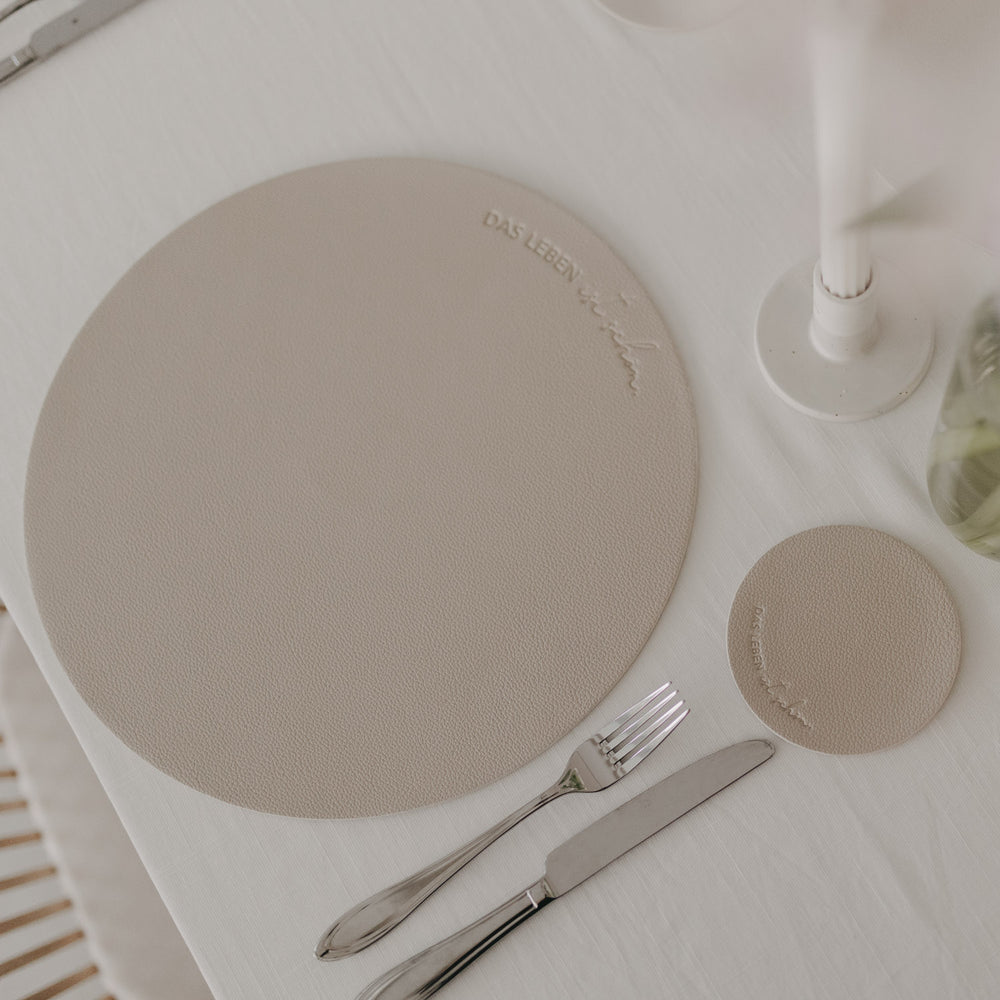 Beiges Platzset mit Untersetzer „Das Leben ist schön“ neben Besteck auf weißer Tischdecke.
