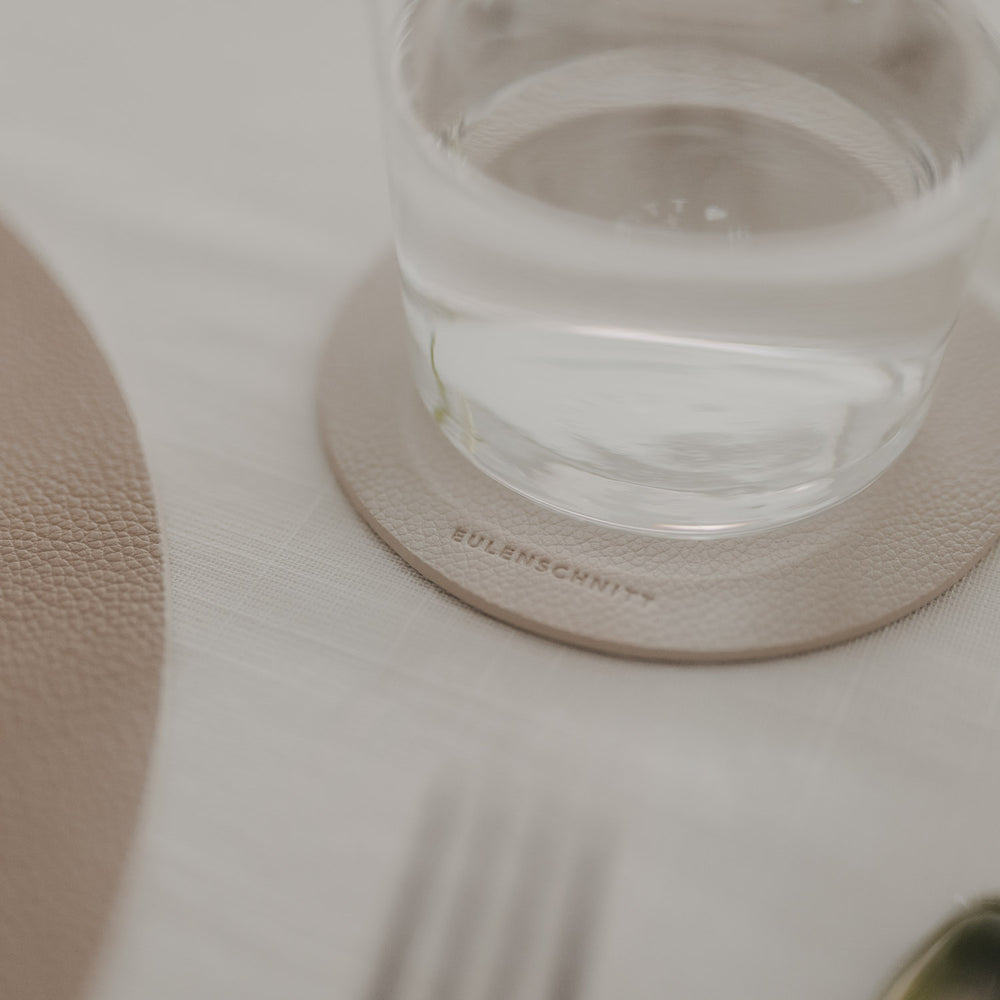 Wasserglas auf beigem Untersetzer mit Eulenschnitt-Prägung auf hell gedecktem Tisch.