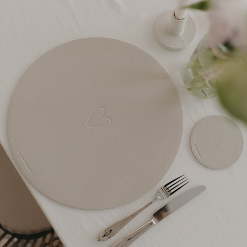 Cremefarbenes Platzset mit Herzprägung und passendem Untersetzer aus Kunstleder auf gedecktem Tisch.