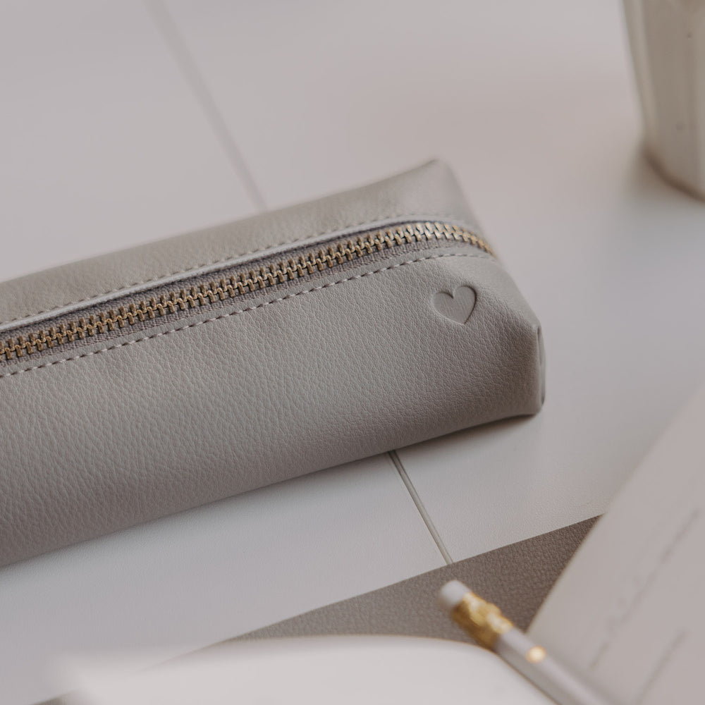 Graue Federtasche mit Herzprägung und goldenem Reißverschluss auf hellem Schreibtisch