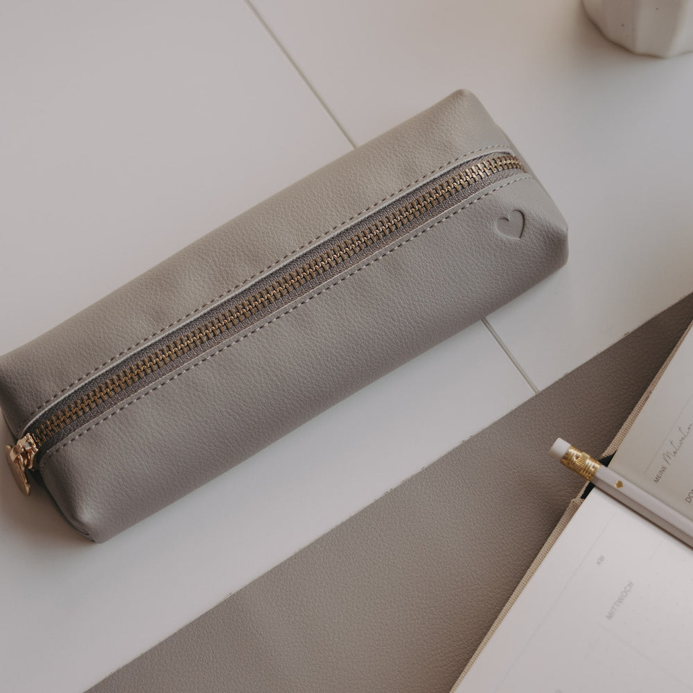 Graue Federtasche mit Herzprägung auf Schreibtisch neben Notizbuch und Bleistift