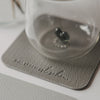 Glas auf grauem quadratischem Untersetzer in Lederoptik mit geprägtem Schriftzug