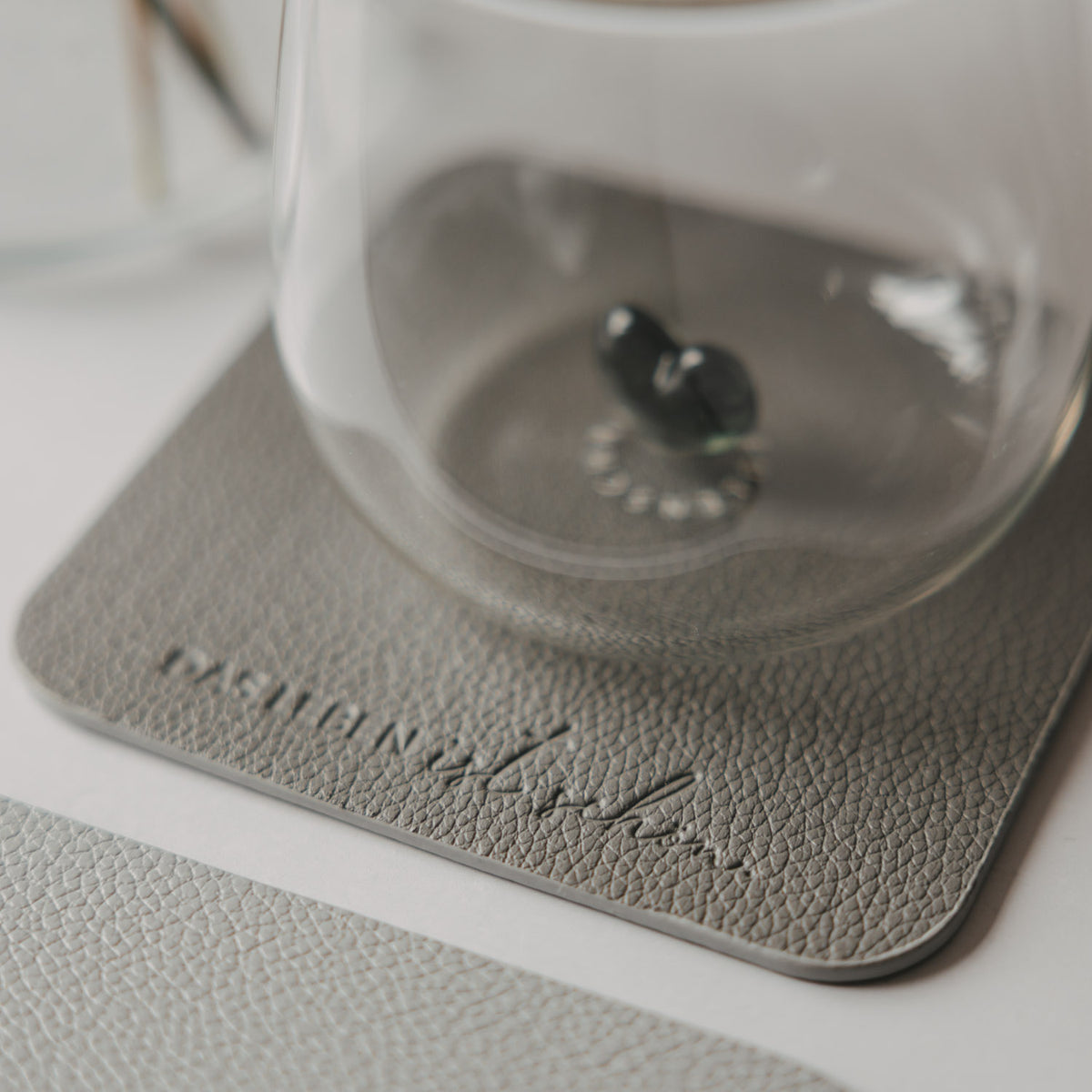 Glas auf grauem quadratischem Untersetzer in Lederoptik mit geprägtem Schriftzug