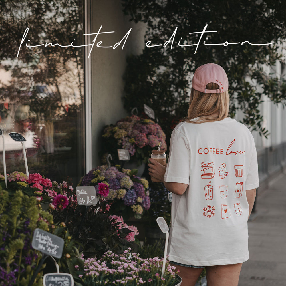 Frau im weißen „Coffee Lover“-T-Shirt und rosa Cap mit Iced Coffee vor einem Blumenladen.