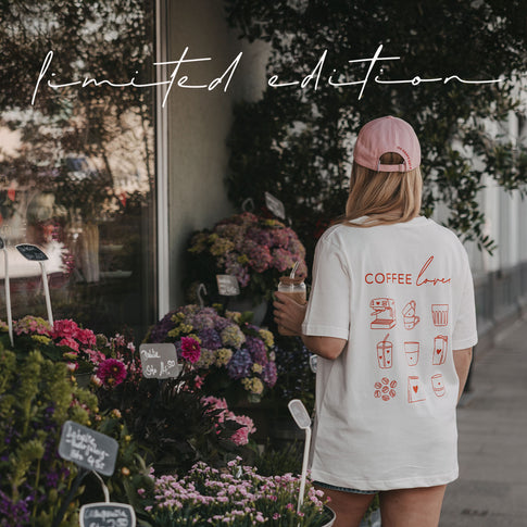 Frau im weißen „Coffee Lover“-T-Shirt und rosa Cap mit Iced Coffee vor einem Blumenladen.