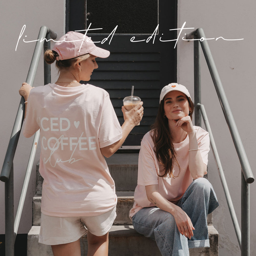 Zwei Frauen in rosafarbenen Iced-Coffee-Shirts sitzen und stehen mit einem Iced Coffee auf einer Treppe.