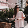 Frau in rosafarbenem Iced-Coffee-T-Shirt mit Sonnenbrille neben einem Fahrrad mit Korb vor einem rosa Gebäude.