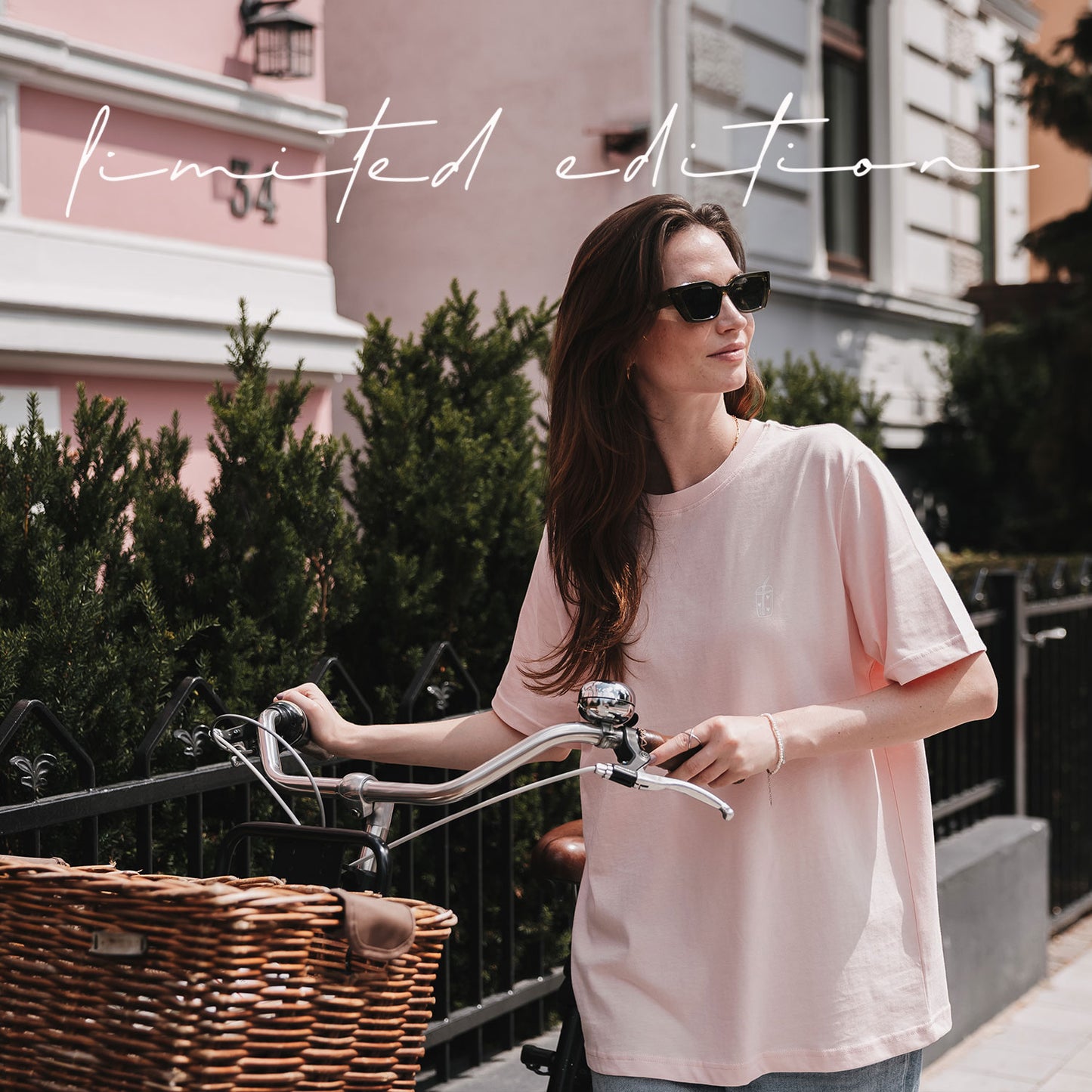 Frau in rosafarbenem Iced-Coffee-T-Shirt mit Sonnenbrille neben einem Fahrrad mit Korb vor einem rosa Gebäude.
