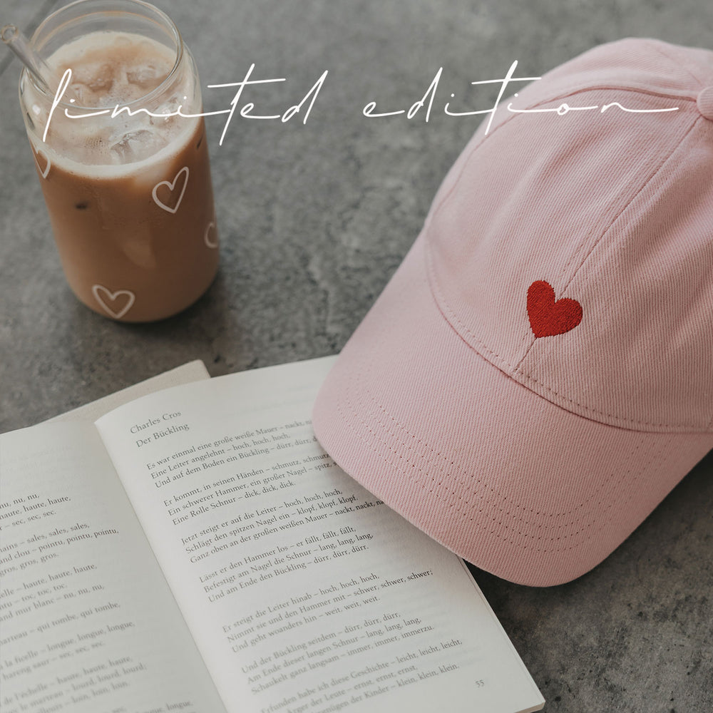 Rosa Cap mit rotem Herz neben Iced Coffee und aufgeschlagenem Buch