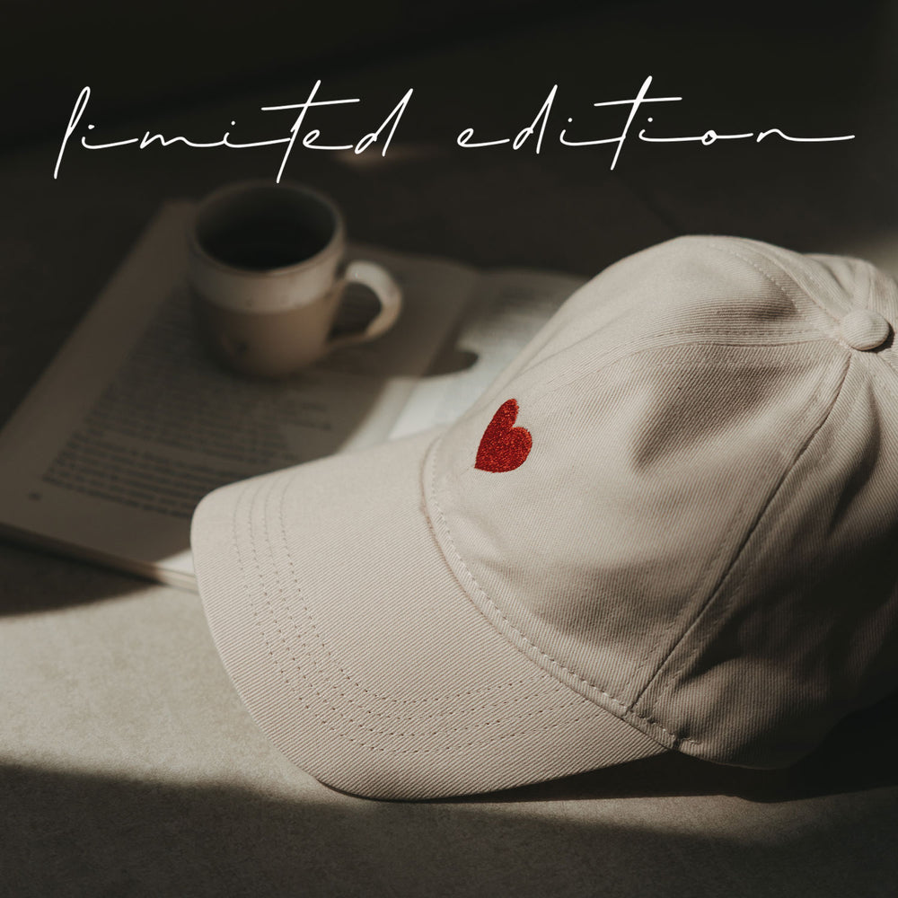 Beige Cap mit roter Herz-Stickerei neben Buch und Kaffeetasse