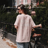 Rückansicht einer Frau im rosafarbenen Oversize-T-Shirt mit weißem „Iced Coffee Club“-Print neben einem Fahrrad.