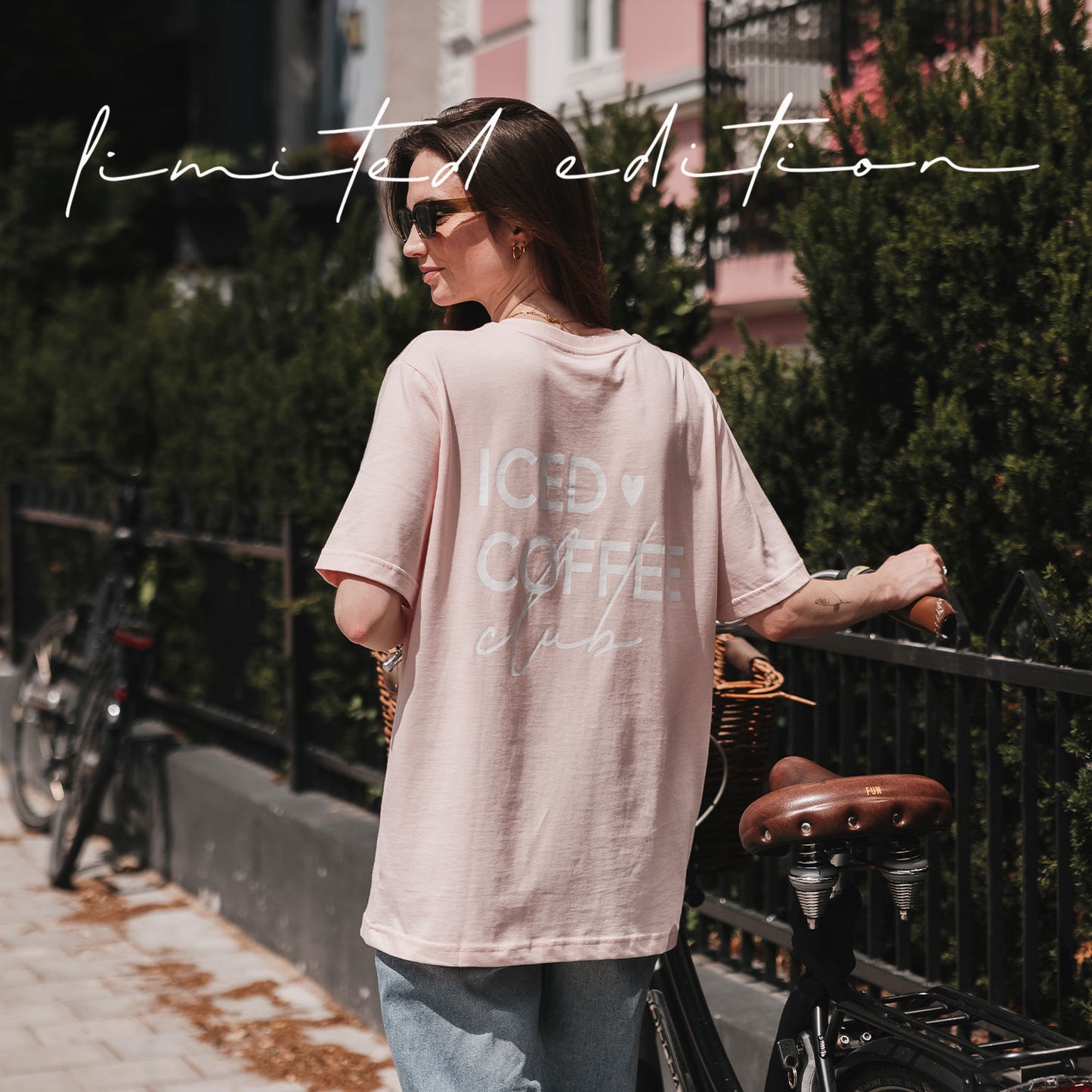 Rückansicht einer Frau im rosafarbenen Oversize-T-Shirt mit weißem „Iced Coffee Club“-Print neben einem Fahrrad.