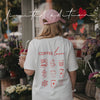Frau mit rosa Cap und weißem Coffee Lover T-Shirt vor einem Blumengeschäft