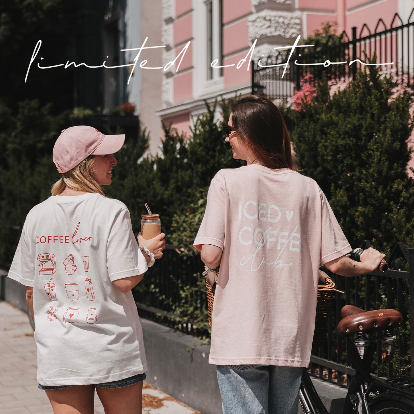Zwei Frauen in weißem „Coffee Lover“- und rosafarbenem „Iced Coffee Club“-T-Shirt stehen mit Iced Coffee und Fahrrad auf der Straße.