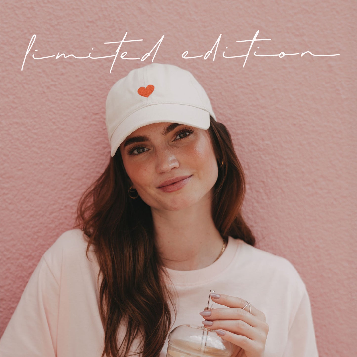 Frau mit beiger Cap mit roter Herz-Stickerei und Iced Coffee vor einer rosa Wand