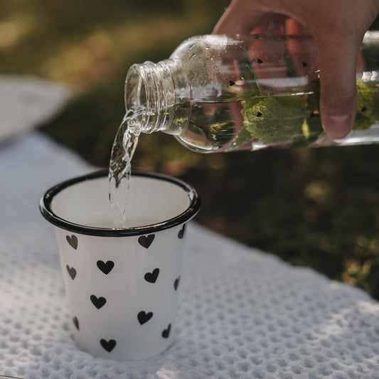 Weißer Emaille-Becher mit schwarzem Herzmuster wird im sommerlichen Picknick-Setting mit Wasser befüllt.
