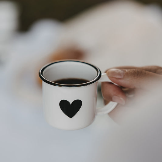 Weiße Emaille-Espressotasse mit schwarzem Herz, gefüllt mit Espresso, wird am Henkel gehalten.