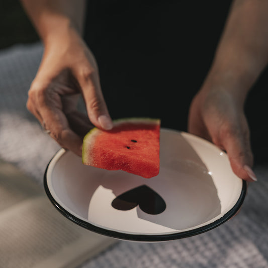 Weißer Emaille-Frühstücksteller mit schwarzem Herz, auf dem eine Wassermelonenscheibe serviert wird.