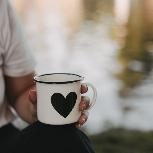 Weiße Emaille-Tasse mit schwarzem Herz wird am See in der Hand gehalten.