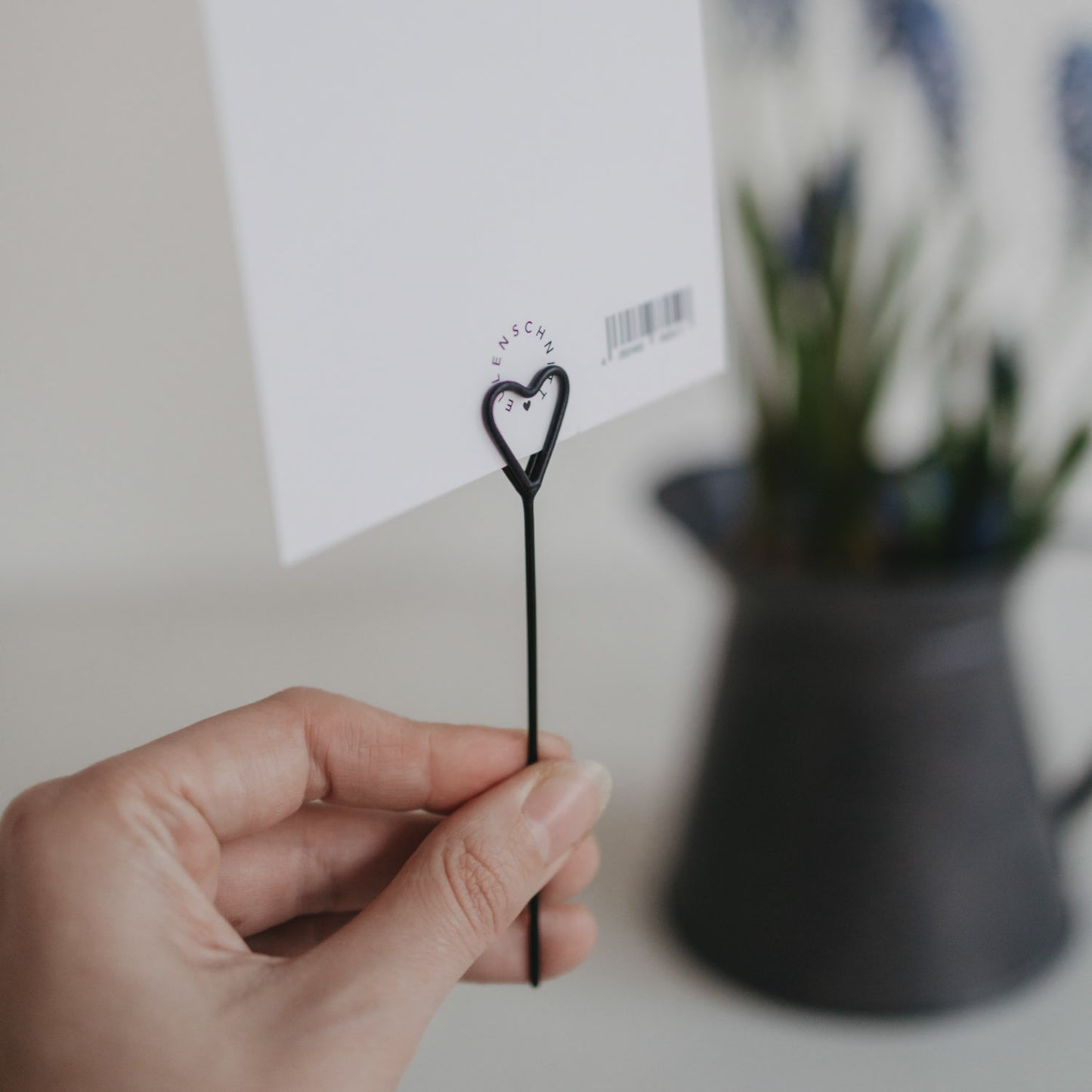Rückseite einer Postkarte im schwarzen Herz-Stecker, gehalten vor unscharfer Vase mit Blumen.