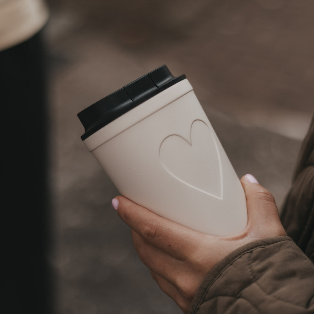 Cremefarbener To-Go-Kaffeebecher mit Herzmotiv und schwarzem Deckel in einer Hand
