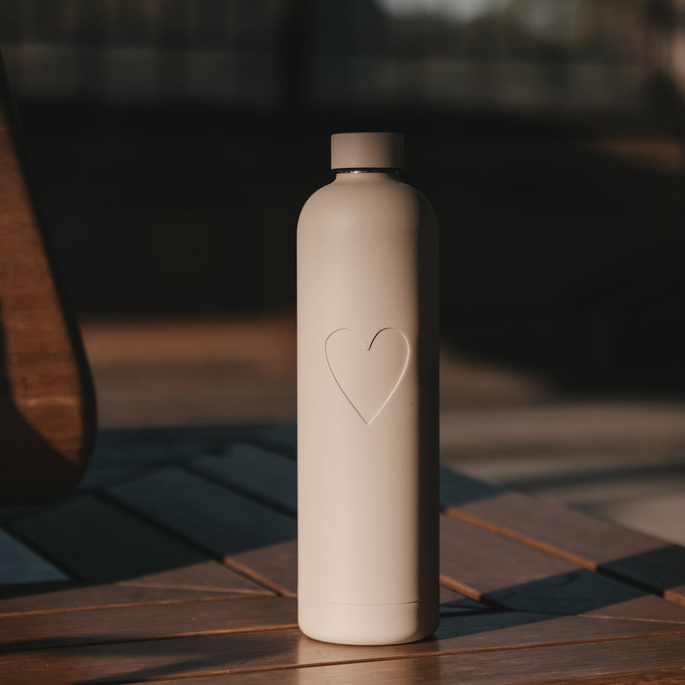 Cremefarbene Trinkflasche mit geprägtem Herzmotiv, 1000 ml und passendem Schraubverschluss auf einem Holztisch.
