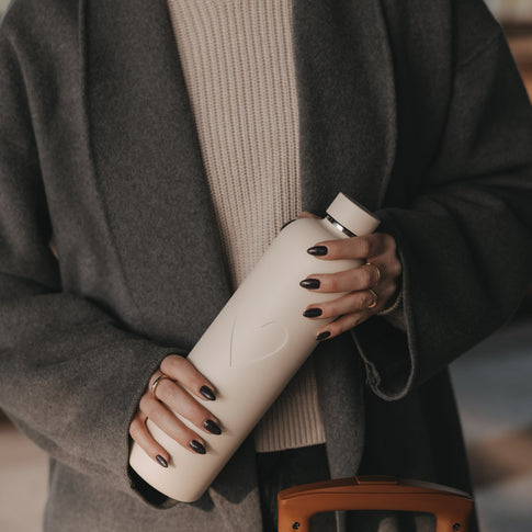 Frau im grauen Mantel hält eine cremefarbene 1000 ml Trinkflasche mit geprägtem Herzmotiv in beiden Händen.