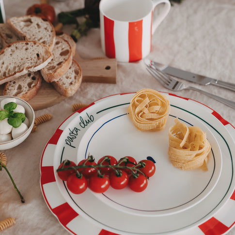Platzteller mit rotem Streifenrand, Speiseteller mit grünem Pasta-Club-Schriftzug und Frühstücksteller mit blauem Herz aufeinander gestapelt, kombiniert mit Pasta, Tomaten und passendem Becher im mediterranen Setting.