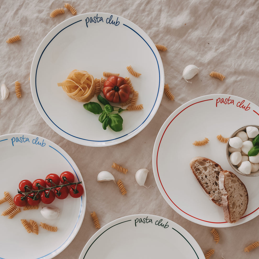 Speiseteller „pasta club“ in verschiedenen Farben (blau, rot, grün, hellblau), dekoriert mit Pasta, Tomaten und Basilikum, auf hellem Leinentisch.