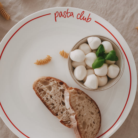 Speiseteller „pasta club“ mit rotem Rand, angerichtet mit Brot, Mozzarella, Basilikum und Pasta auf hellem Leinentisch.