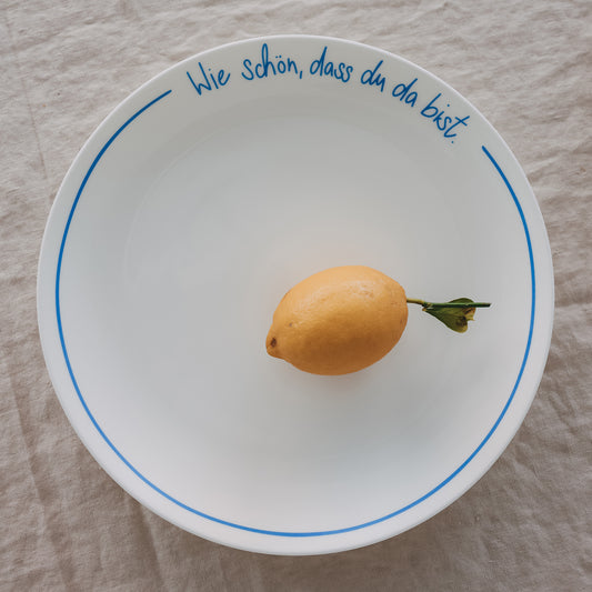 Weißer Platzteller mit hellblauer Schrift „Wie schön, dass du da bist“ mit Zitrone, auf hellem Leinentisch.
