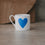 Kleine weiße Tasse mit großem hellblauen Herz frontal auf heller Steinfläche, weicher Hintergrund.