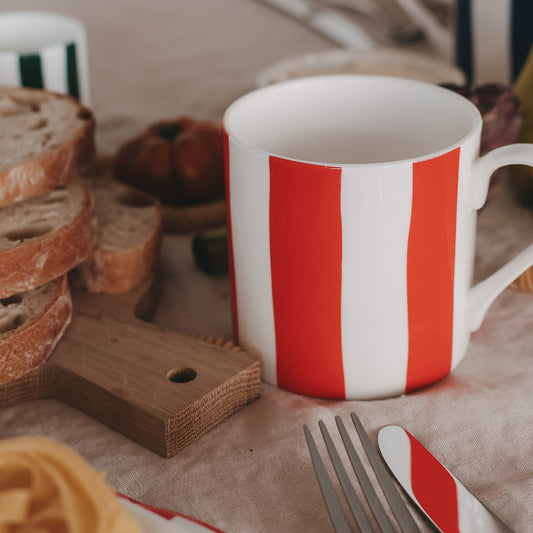 Porzellantasse mit roten und weißen Streifen neben Brot, Holzbrett und Besteck auf gedecktem Tisch