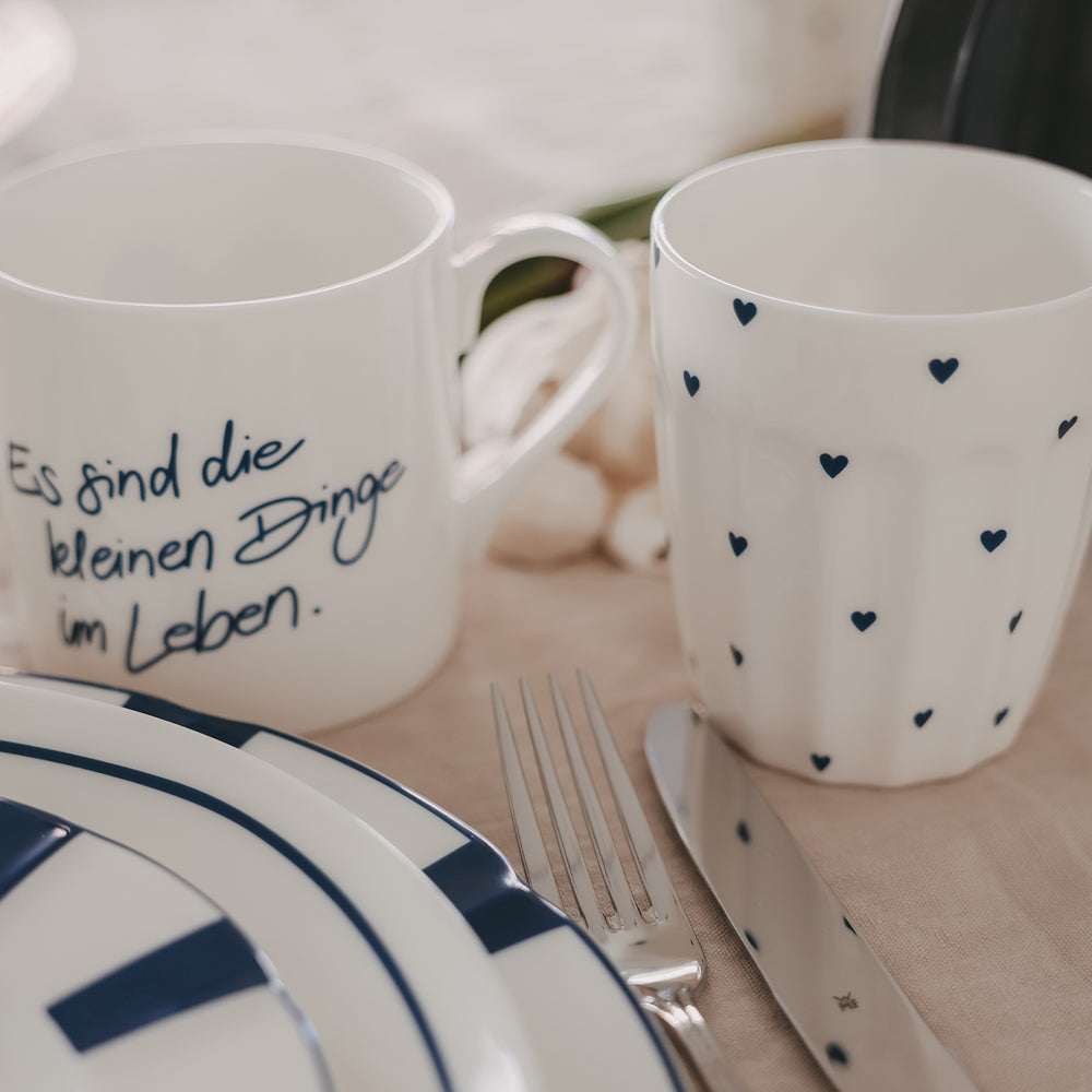 Keramikbecher mit kleinen blauen Herzen auf gedecktem Tisch, neben Geschirr und Besteck, gemütliche Szene