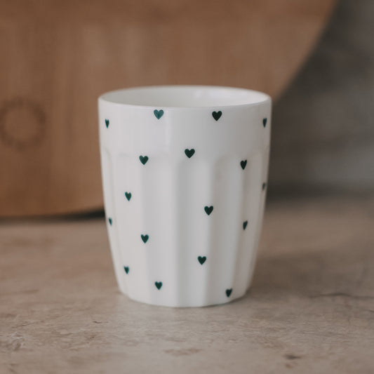 Weißer Keramikbecher mit kleinen grünen Herzen auf Tisch, minimalistisch und handgemacht