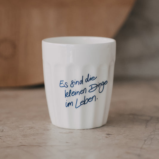 Weißer Keramikbecher mit Schriftzug „Es sind die kleinen Dinge im Leben“ in Blau auf hellem Untergrund