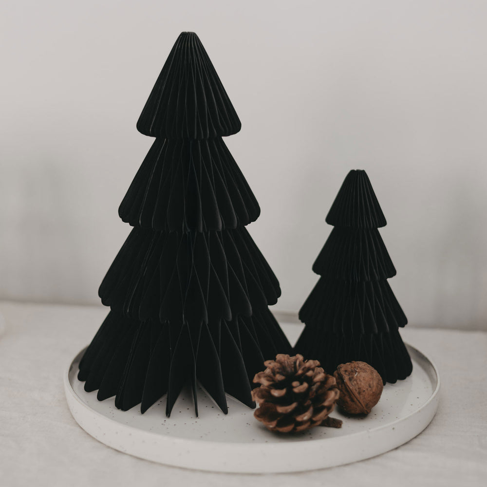 Dekoratives Weihnachtsarrangement mit zwei schwarzen Papiertannen in verschiedenen Größen auf einem weißen Steingutteller, ergänzt durch einen Tannenzapfen und eine Walnuss.