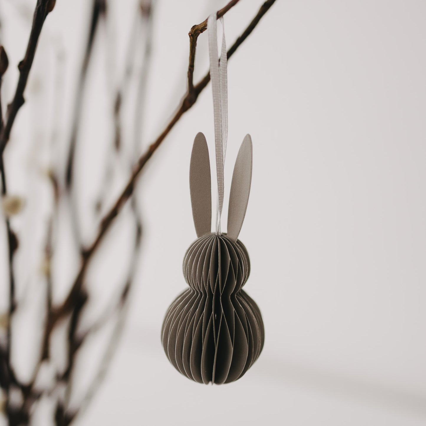 Grauer Papieranhänger in Hasenform mit weißen Ohren hängt an einem Zweig.