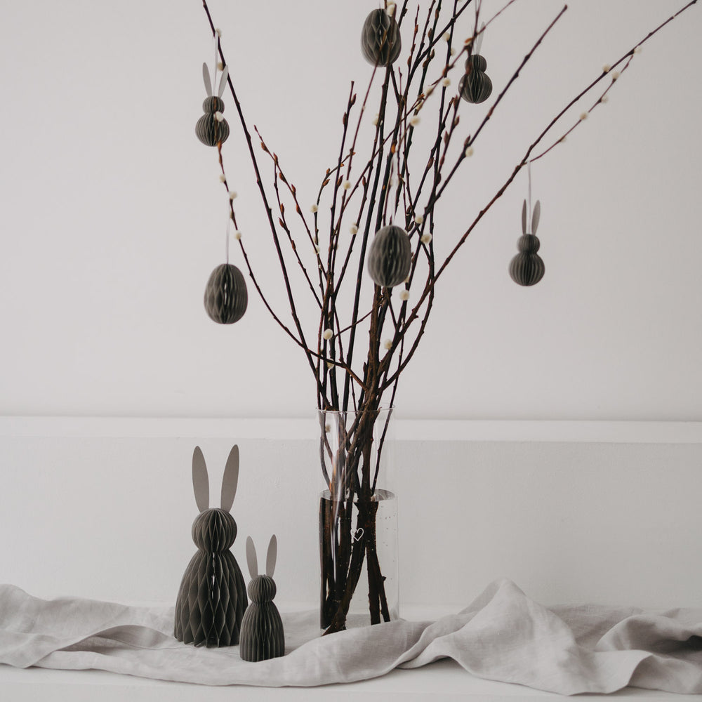 Graue Osteranhänger aus Papier hängen an Zweigen in einer Glasvase, dekoriert mit Hasenfiguren.