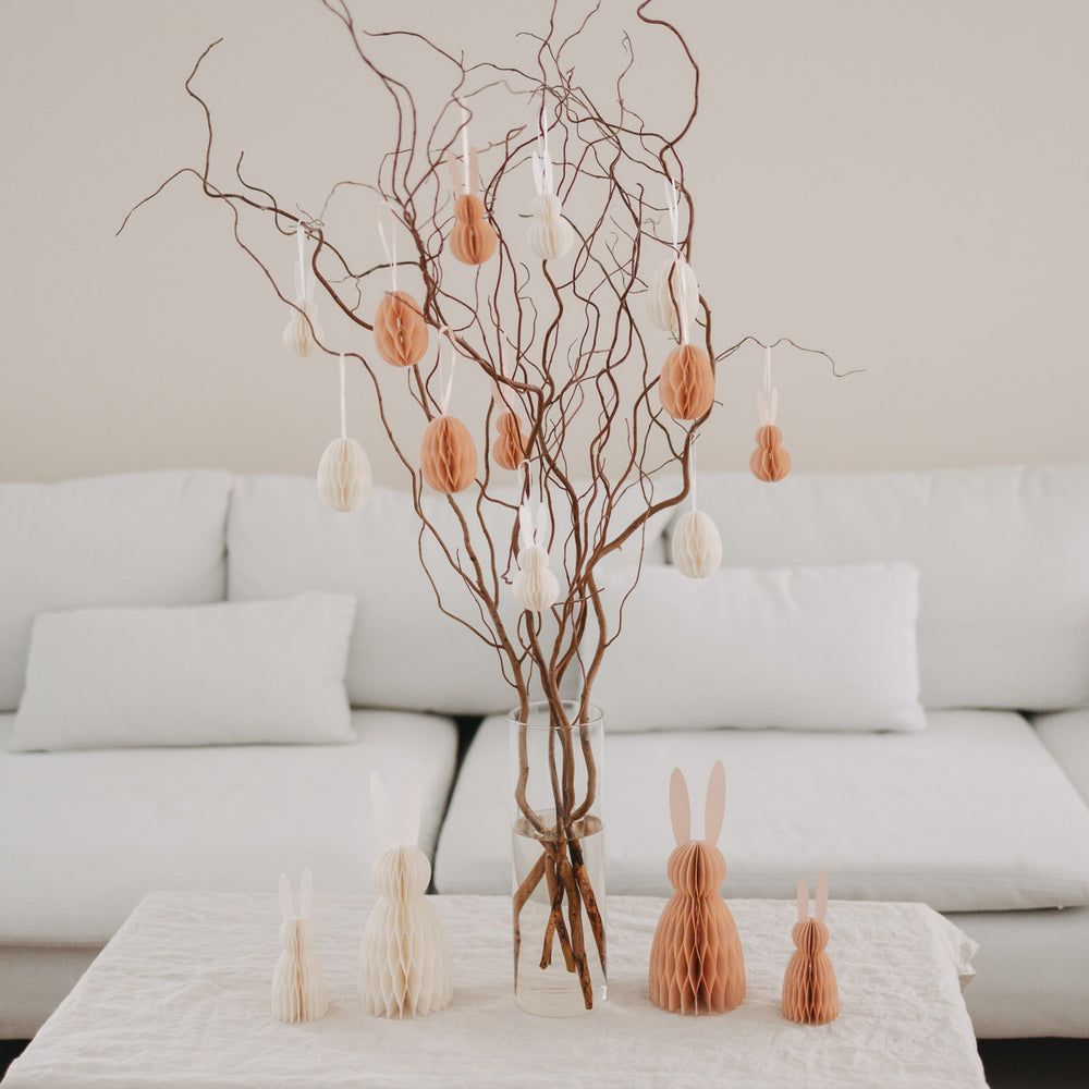 Pfirsichfarbene Papierhasen als Osterdeko neben Osterzweigen und Anhängern im Wohnzimmer.