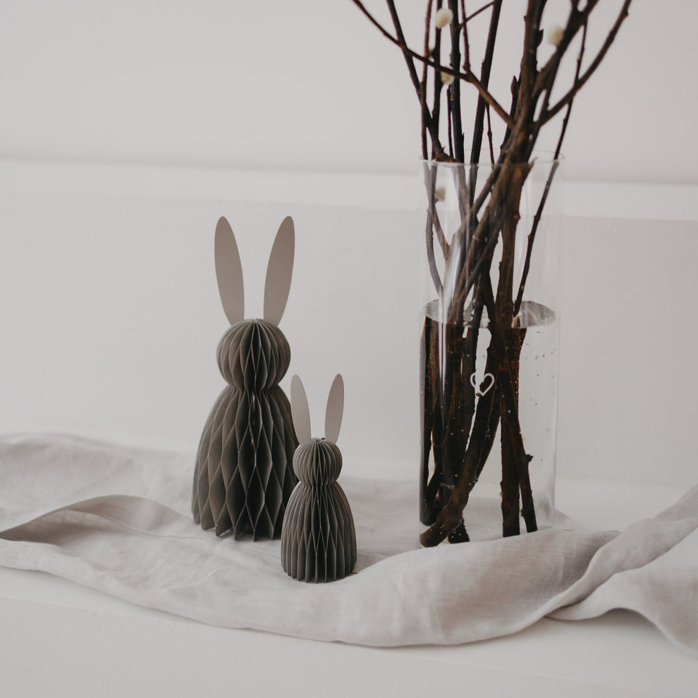 Papierhasen in Grau als Deko, zwei Größen, auf Stofftuch neben Vase mit Zweigen arrangiert.