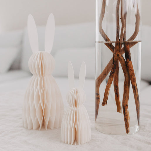 Zwei beige Papierhasen im Honeycomb-Design als Deko, stehend auf Stoff neben Glasvase mit Ästen.